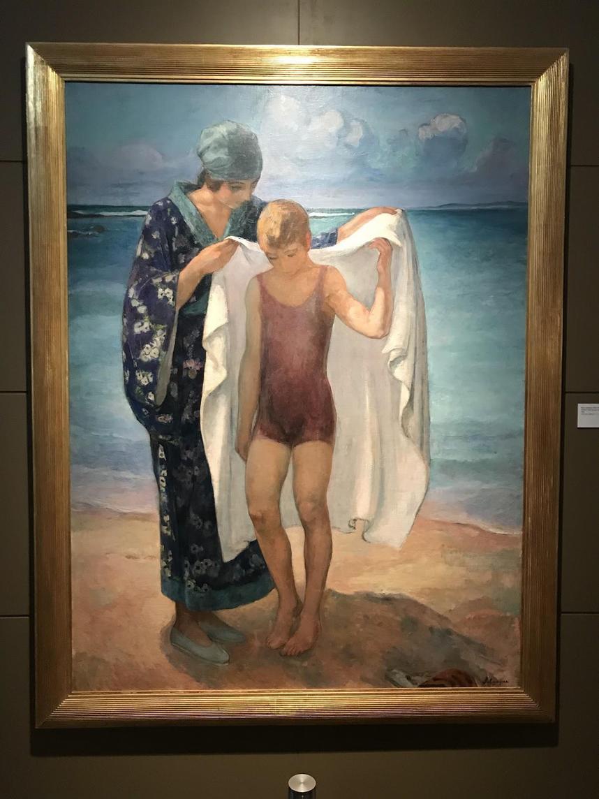 Henri Lebasque - Deniz Banyosu Ön planda, kumda, koyu mavi renkte üstünde beyaz çiçek desenleri olan bornoz ve açık mavi renkte ayakkabılar ve aynı renkte bone giyen bir kadın görüyoruz.