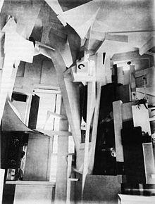 Ayça Alper Akçay Merzbau nun çekirdeğini oluşturan heykellerden ilki sayılan Istırap (Leiden, 1917) adlı çalışma, ahşap, mukavva, hurda demir levhalar, kırık mobilya parçaları, tablo çerçeveleri gibi